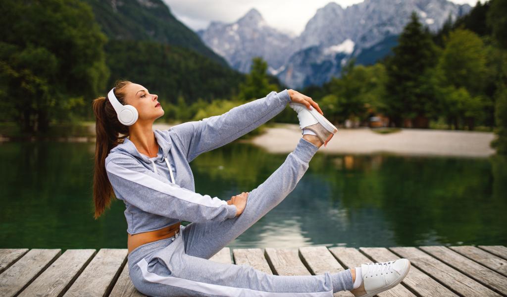 Девушка в наушниках занимается гимнастикой на мосту у озера