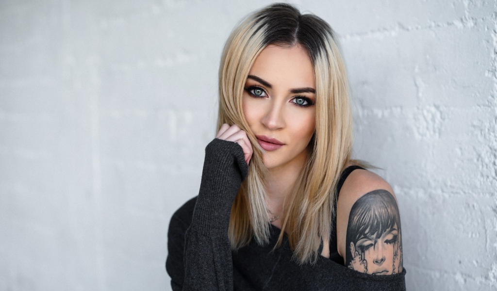 Стильная блондинка с татуировкой на плече на фоне стены