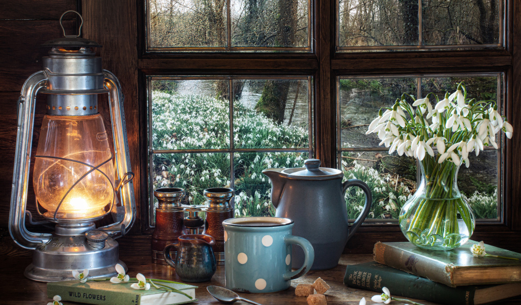Керосиновая лампа на столе с кружкой, книгами и букетом подснежников у окна