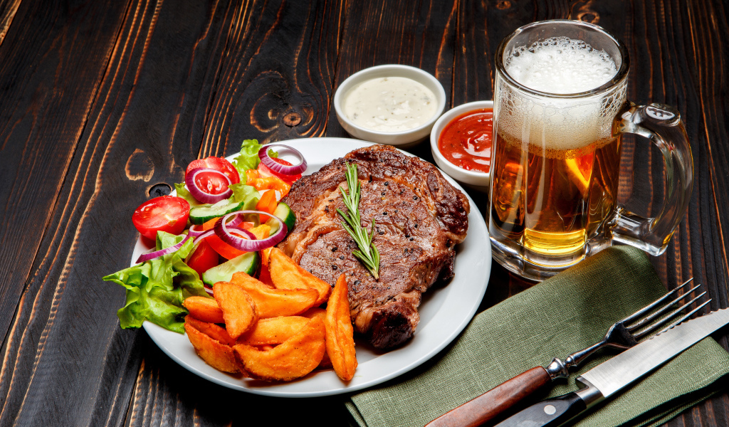 Кусок мяса на тарелке с картофелем и салатом на столе с соусом и пивом