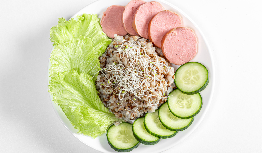 Гречка на тарелке с колбасой, огурцами и листьями салата