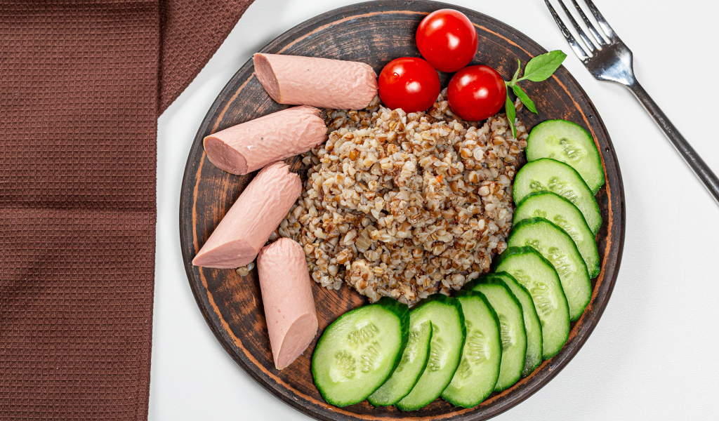 Гречка на тарелке с помидорами, огурцами и сосисками