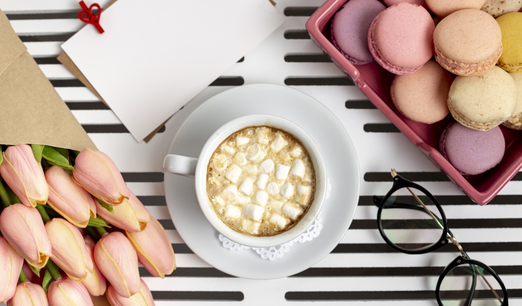 Чашка какао с маршмеллоу на столе с букетом тюльпанов, десертом макарун и очками