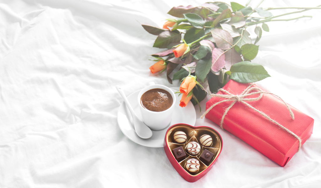 Чашка кофе на белой кровати с конфетами, подарком и букетом роз