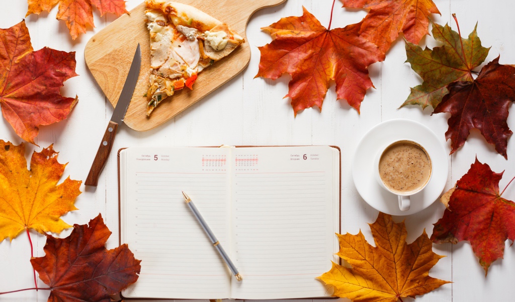 Ежедневник на столе с желтыми листьями, кофе и пиццей 