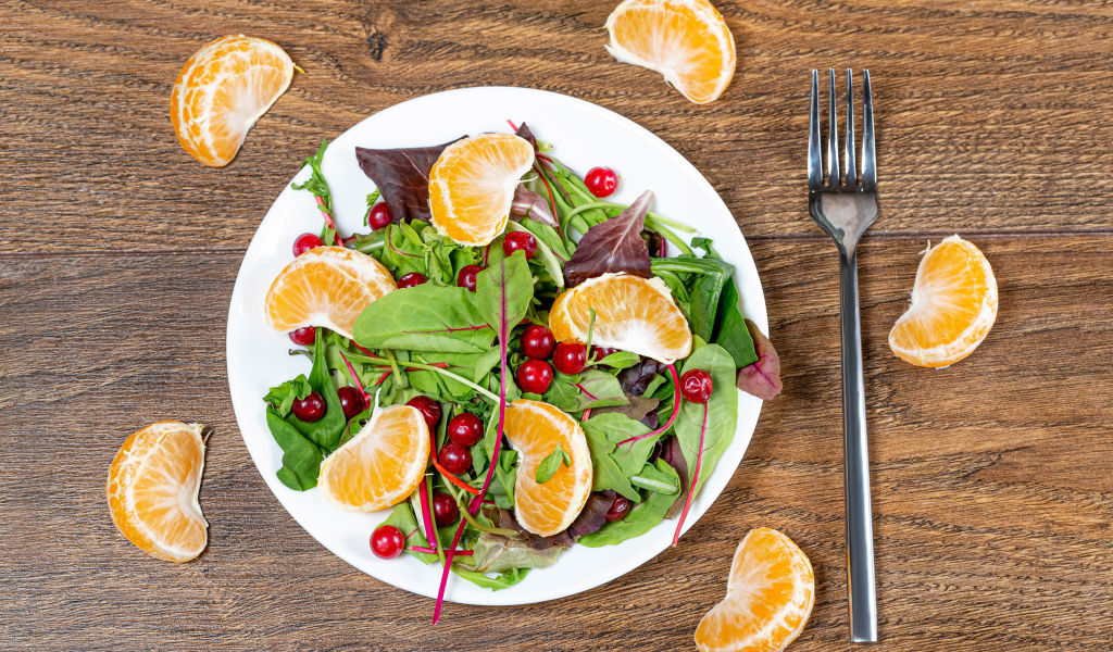 Салат из зелени и мандарина на белой тарелке на столе с вилкой