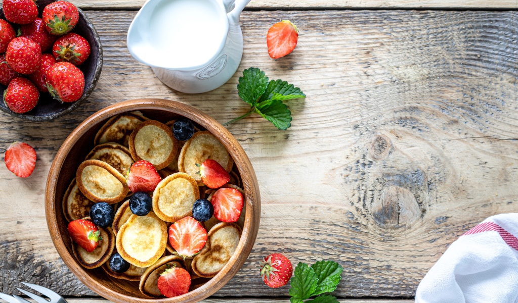Маленькие оладьи в миске с клубникой и черникой на столе с молоком 