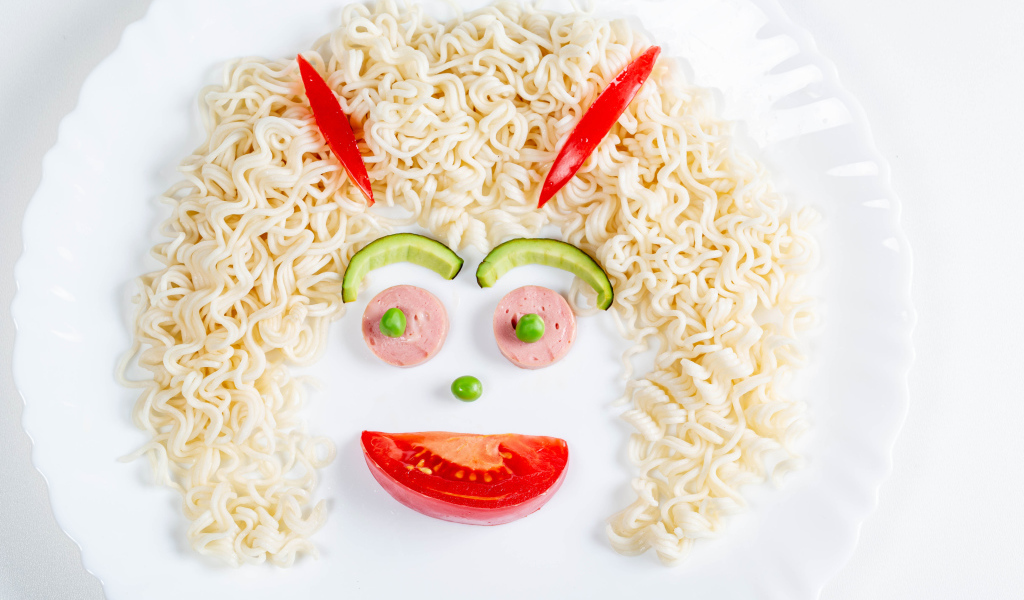 Лицо из вермишели с овощами и колбасой на белой тарелке 