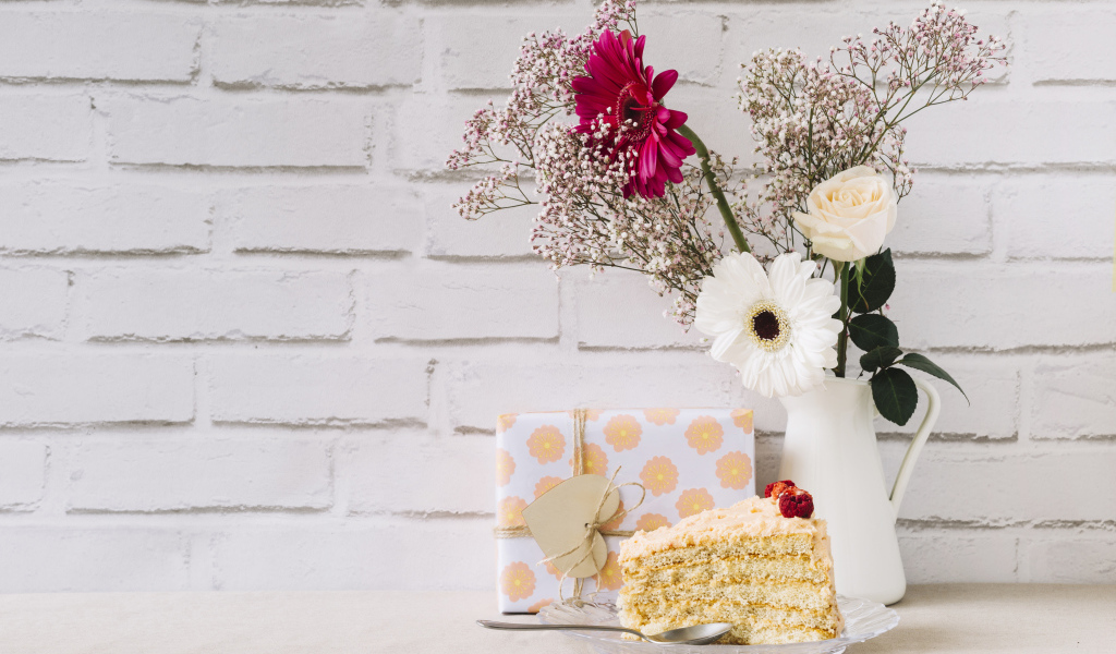 Кусок торта, подарок и букет на столе на фоне стены 