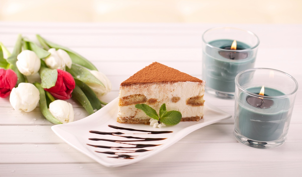 Кусок тирамису на столе с зажженными свечами и букетом тюльпанов