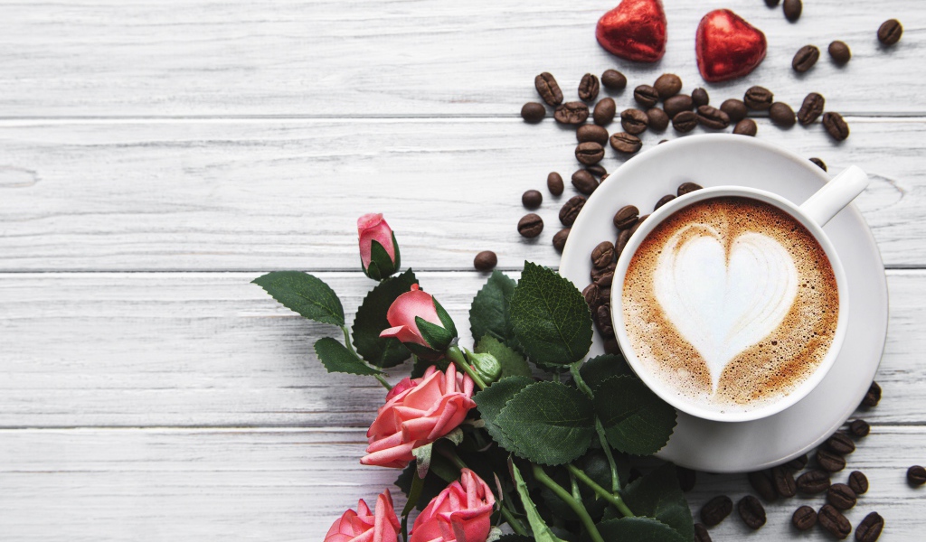 Чашка кофе с рисунком на пене на столе с букетом роз, зернами и конфетами