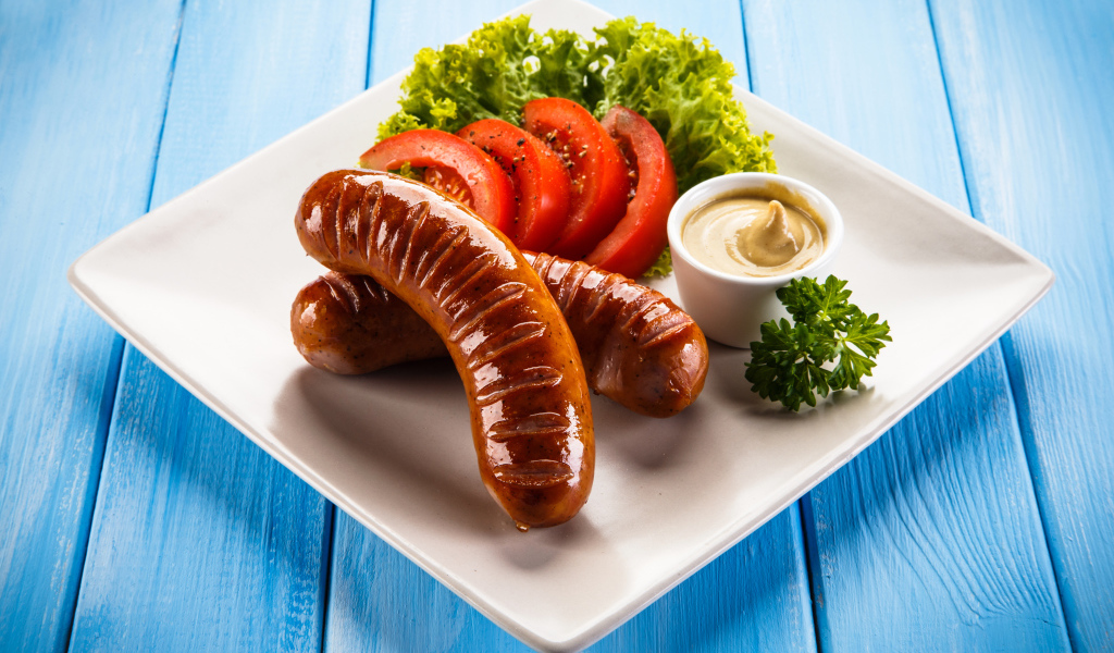 Две жареные колбаски на тарелке с помидорами, горчицей и листьями салата 