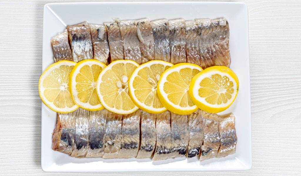 Селедка нарезанная кусочками на белой тарелке с лимоном 