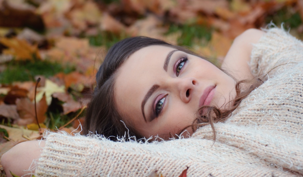 Красивая девушка в теплом свитере лежит на сухой листве 