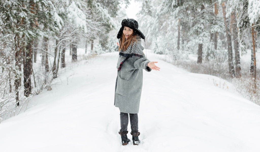 Девушка в пальто и теплой шапке гуляет в заснеженном лесу 