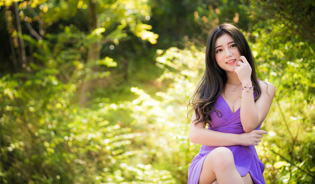 Улыбающаяся девушка азиатка в фиолетовом платье сидит в парке 