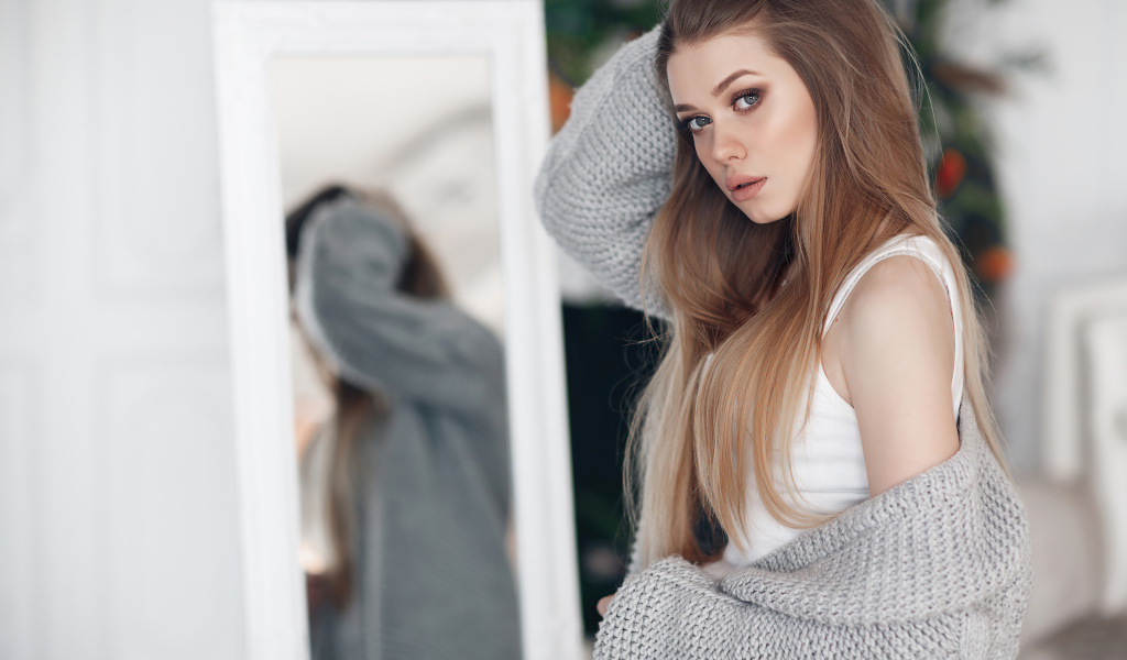 Молодая девушка в серой кофте стоит у зеркала 