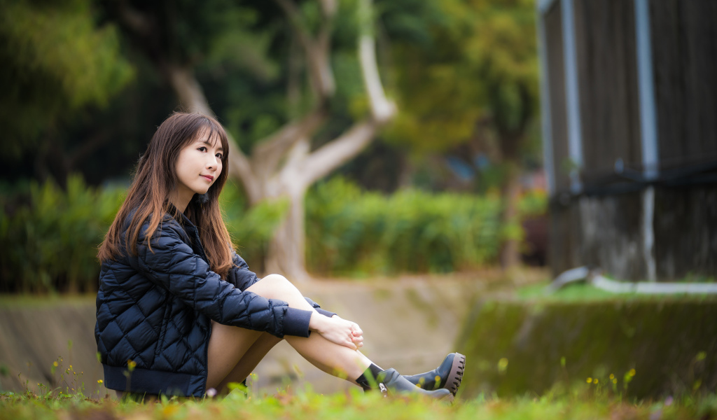 Девушка азиатка в черной куртке сидит на траве