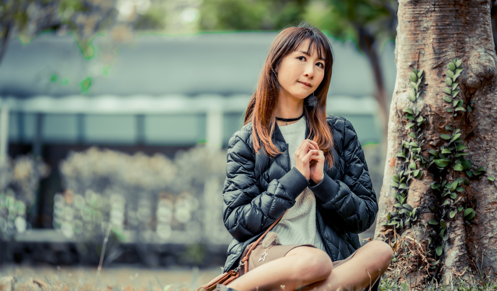 Девушка азиатка в черной куртке сидит под деревом
