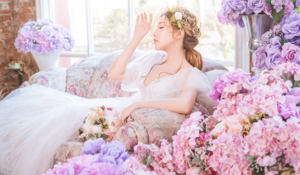 Девушка азиатка в белом платье сидит на диване в комнате с цветами