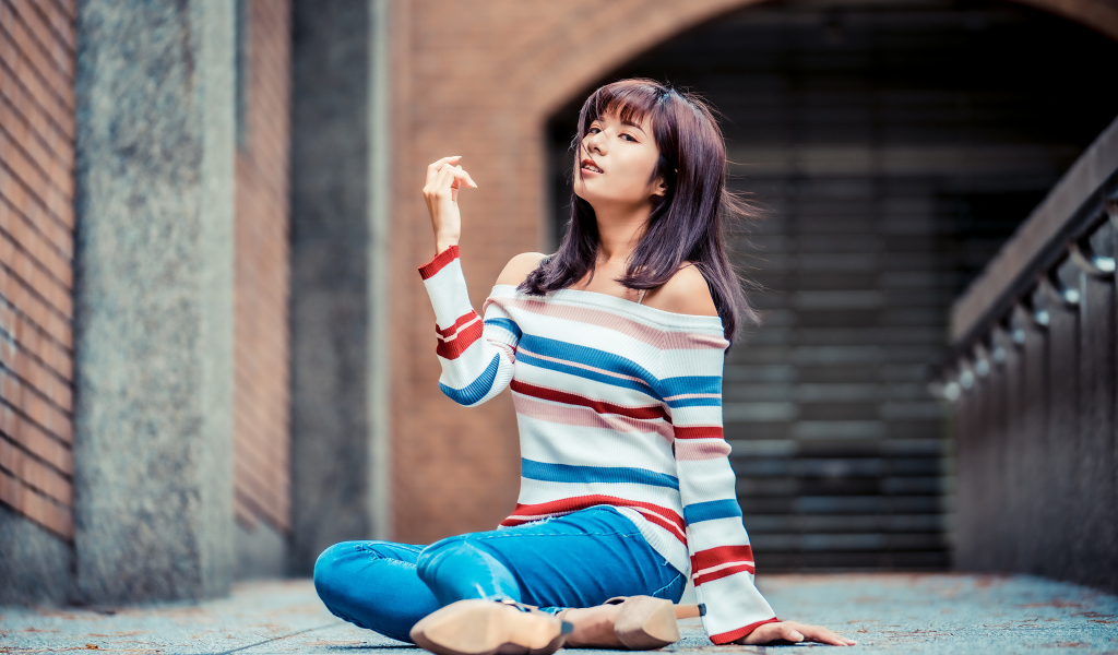 Девушка азиатка в джинсах и полосатом свитере сидит на асфальте