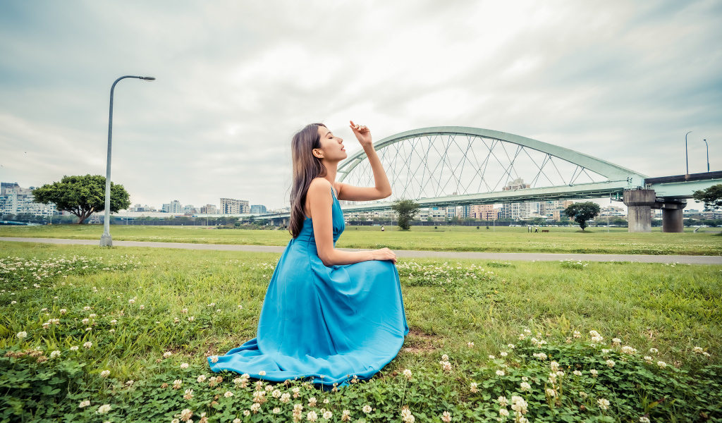 Красивая девушка азиатка в голубом платье  сидит на траве на фоне моста