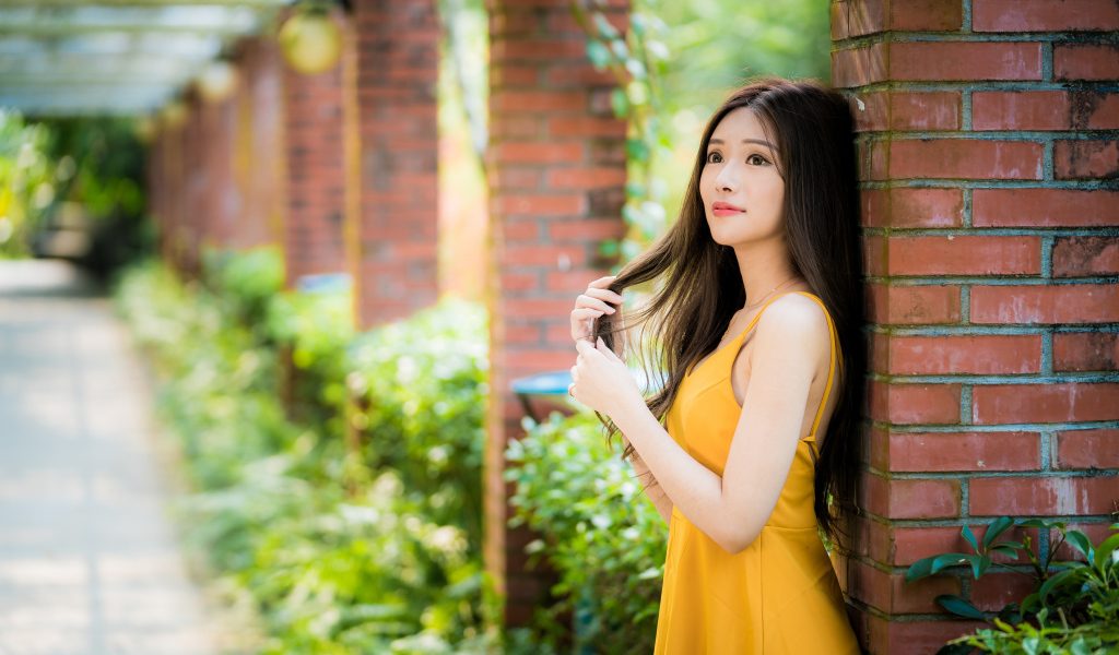 Красивая азиатка в желтом платье стоит у кирпичной колонны 