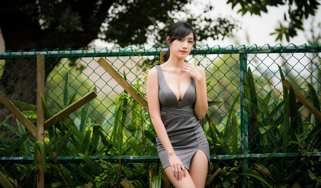 Красивая азиатка в сером платье стоит у зеленой сетки 