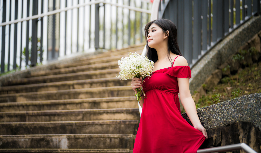 Красивая азиатка в красном платье стоит на ступеньках с букетом ландышей