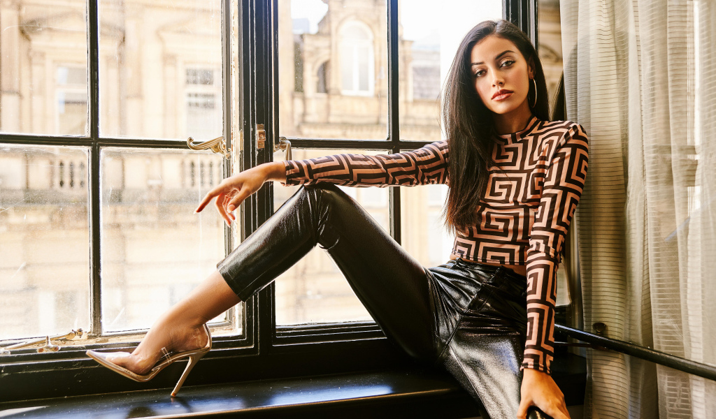 Красивая брюнетка в черных брюках сидит на окне 