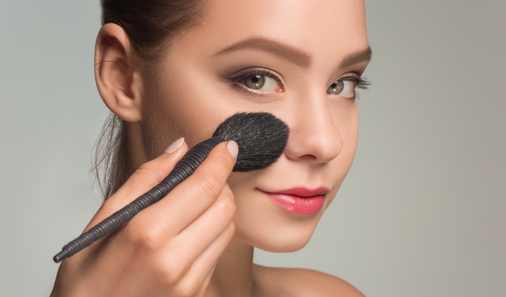 Красивой девушке наносят макияж на лицо кистью 