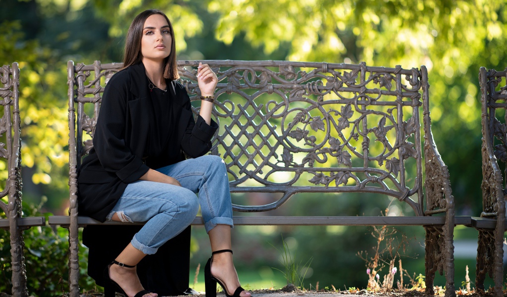Красивая девушка в черном пальто сидит на лавочке в парке