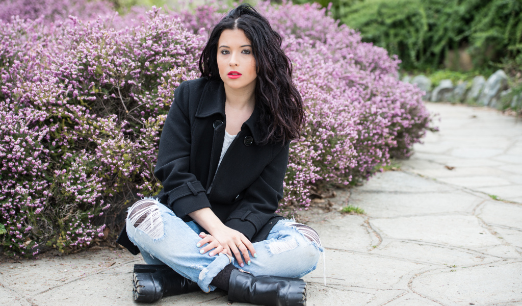 Красивая девушка в черном пальто сидит на земле у кустов