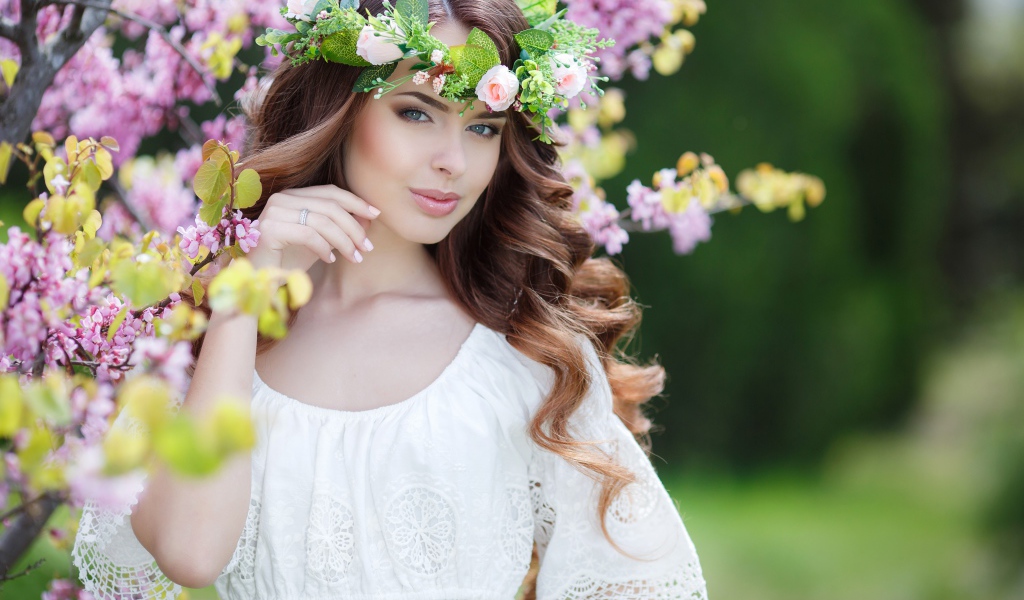 Красивая девушка в белом платье с венком на голове стоит у дерева 