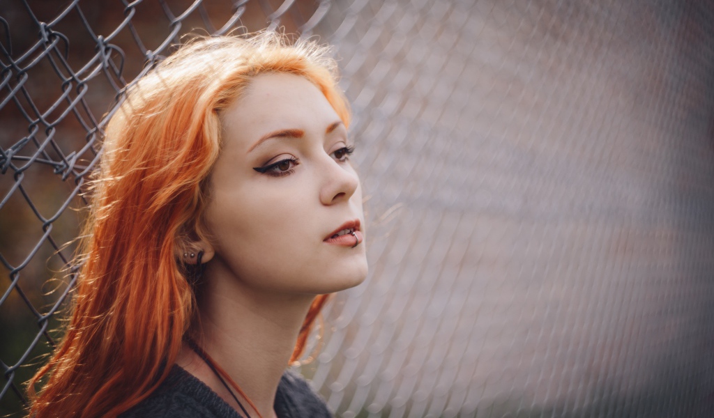 Красивая рыжеволосая девушка с пирсингом у железной сетки 
