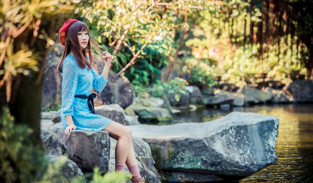 Милая азиатка в берете и синем платье сидит у воды 