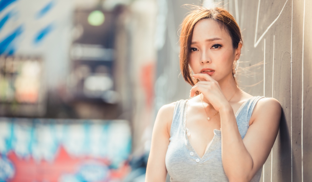 Милая девушка азиатка  с рукой у лица стоит у стены 