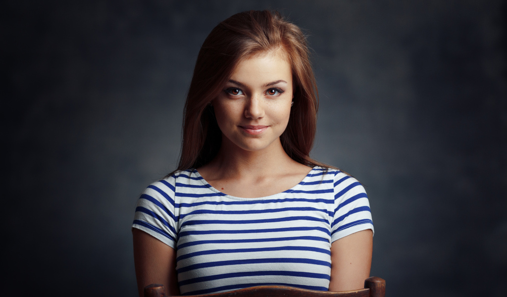 Милая кареглазая девушка в полосатой майке сидит на стуле 