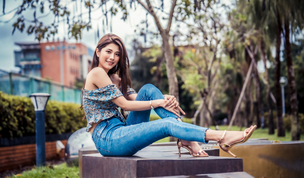Улыбающаяся азиатка в джинсах сидит в парке