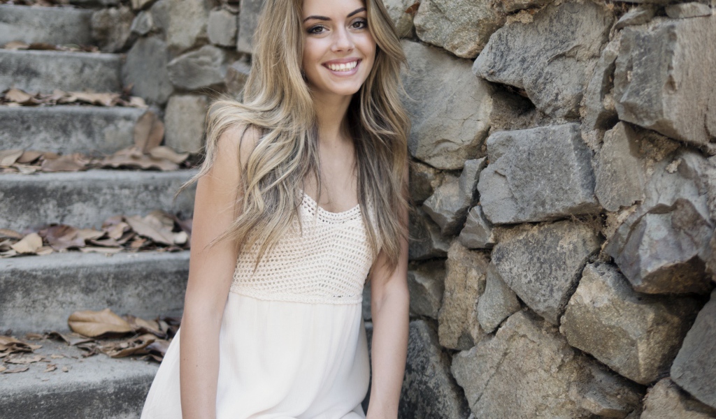 Улыбающаяся  девушка в белом платье стоит на ступеньках у стены