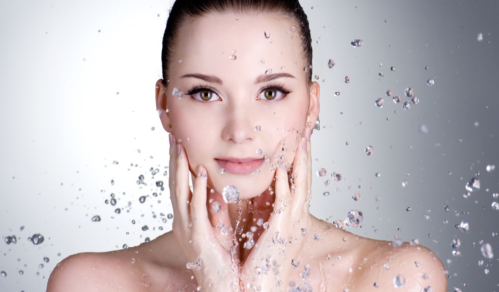 Брызги воды летят на красивое лицо молодой девушки на сером фоне