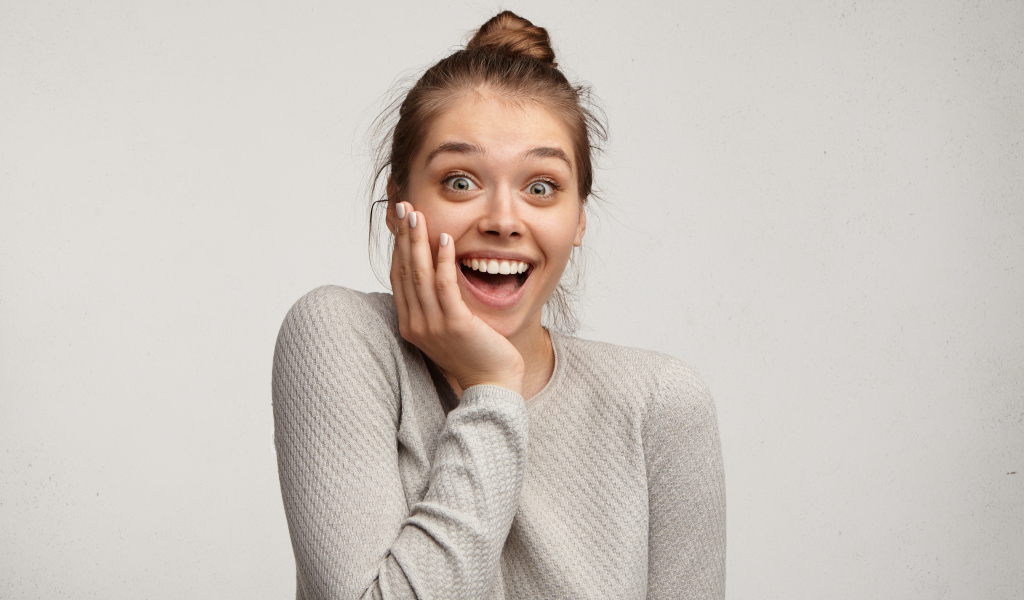 Удивленная девушка с рукой у лица на сером фоне 