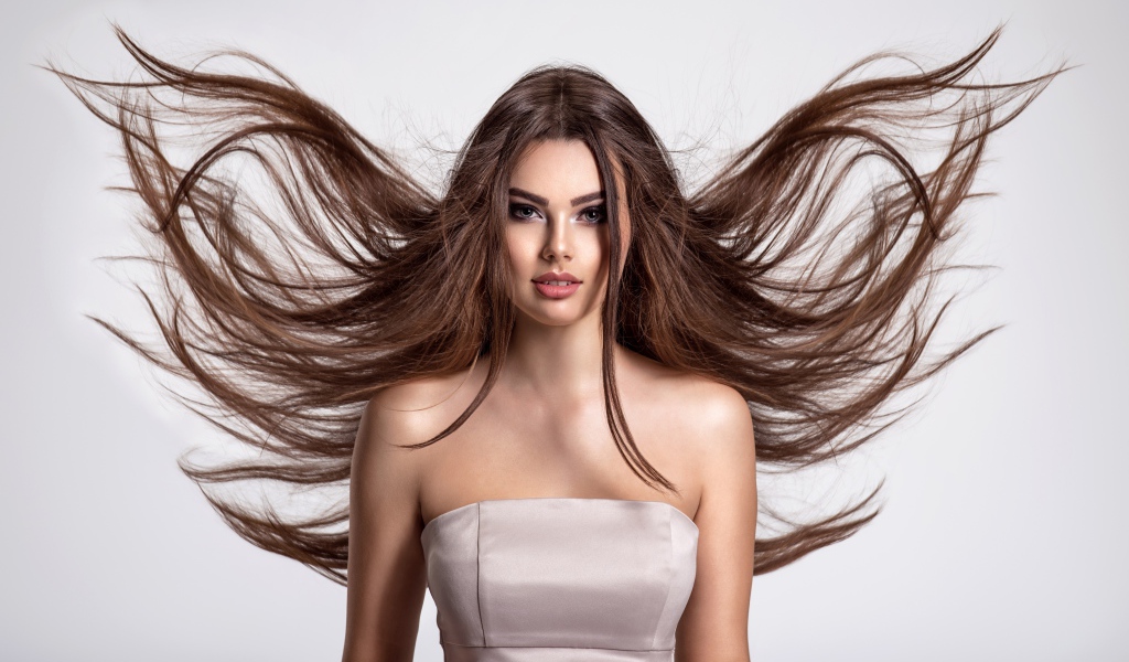 Ветер развивает волосы красивой девушке шатенке на сером фоне