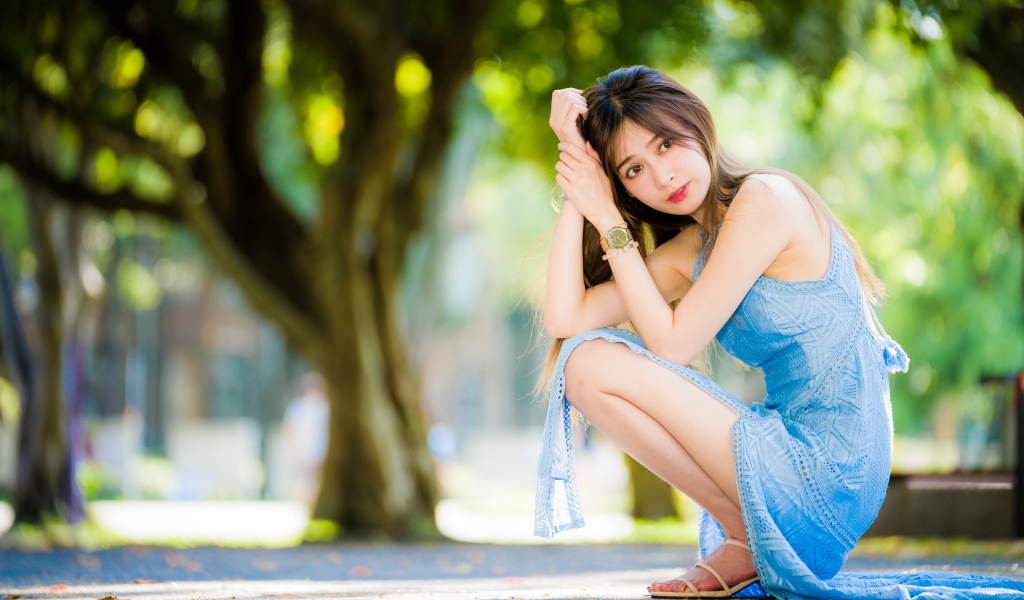 Молодая девушка азиатка в голубом платье сидит на корточках в парке 