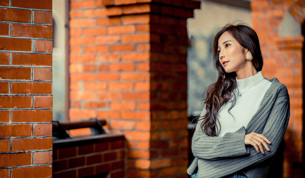Молодая девушка азиатка в серой кофте у кирпичной стены 