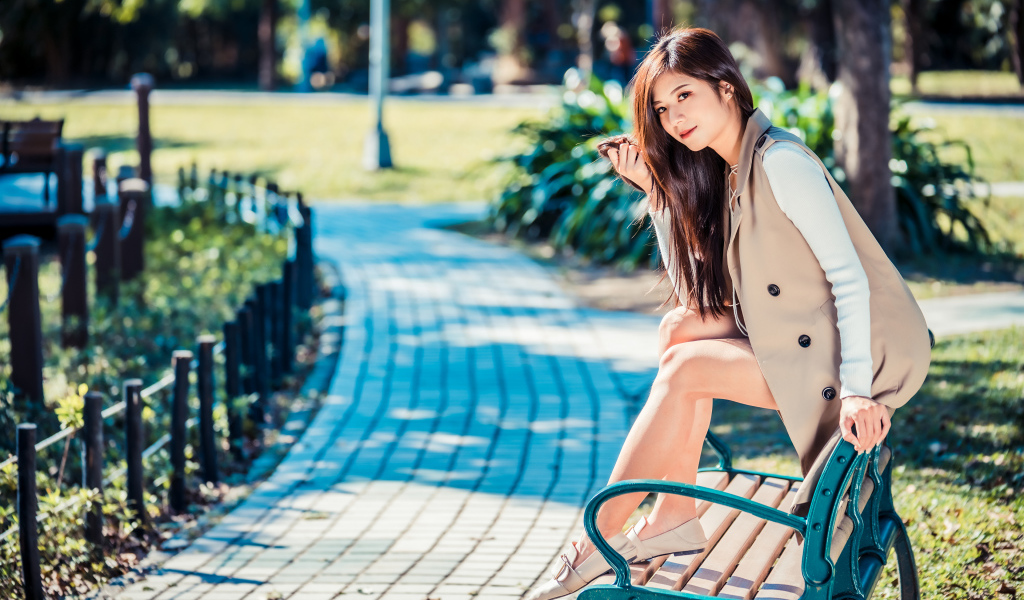 Молодая девушка азиатка сидит в пальто на лавке в парке 