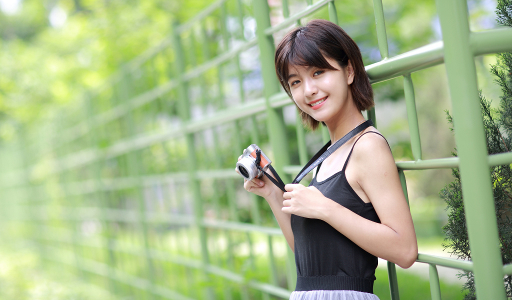 Молодая улыбающаяся азиатка с фотоаппаратом у забора 