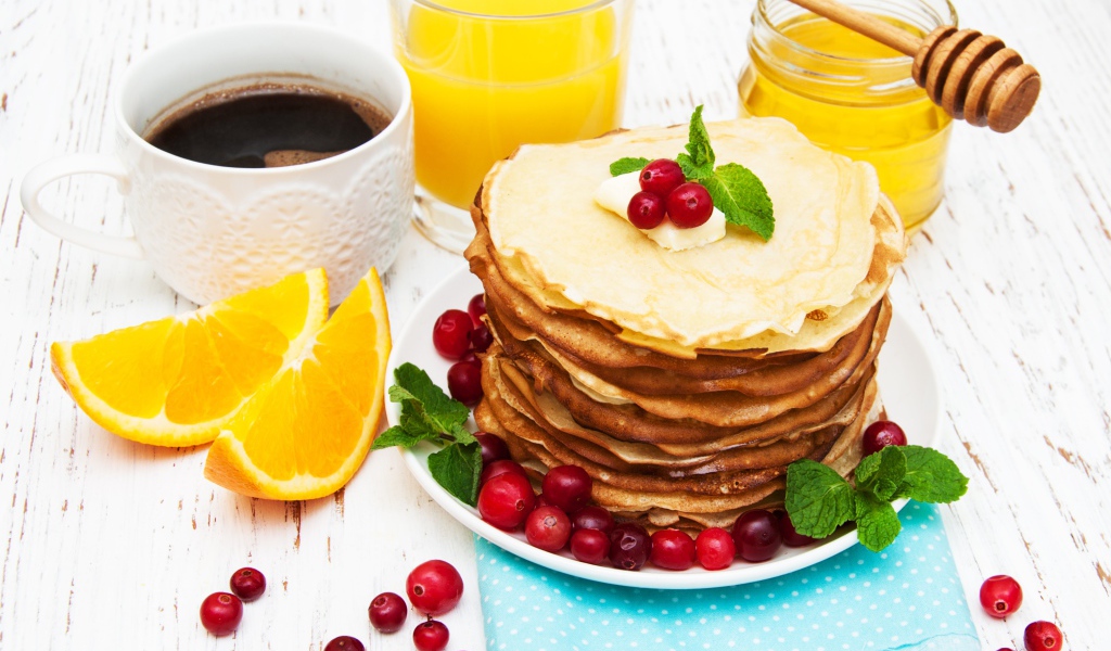Тонкие румяные блинчики на столе с ягодами клюквы, апельсинами, медом и кофе