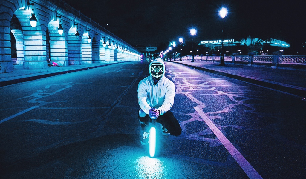 Парень в светящейся маске сидит с неоновой лампой на дороге