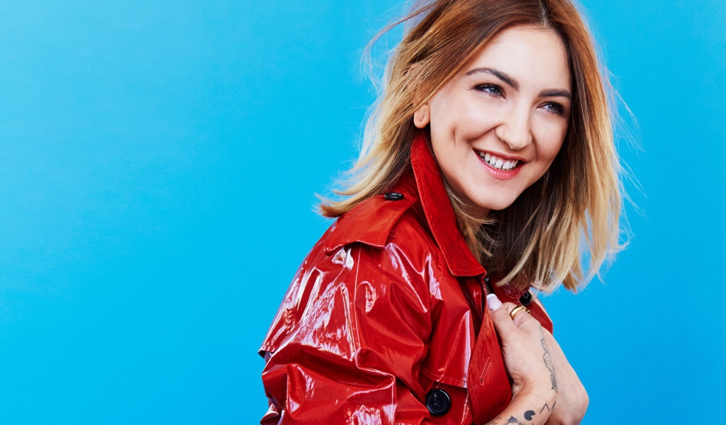 Улыбающаяся певица Джулия Майклз в красной куртке на голубом фоне